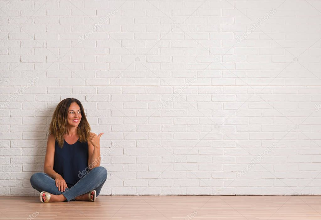 Middle age hispanic woman sitting on the floor over white brick wall smiling with happy face looking and pointing to the side with thumb up.