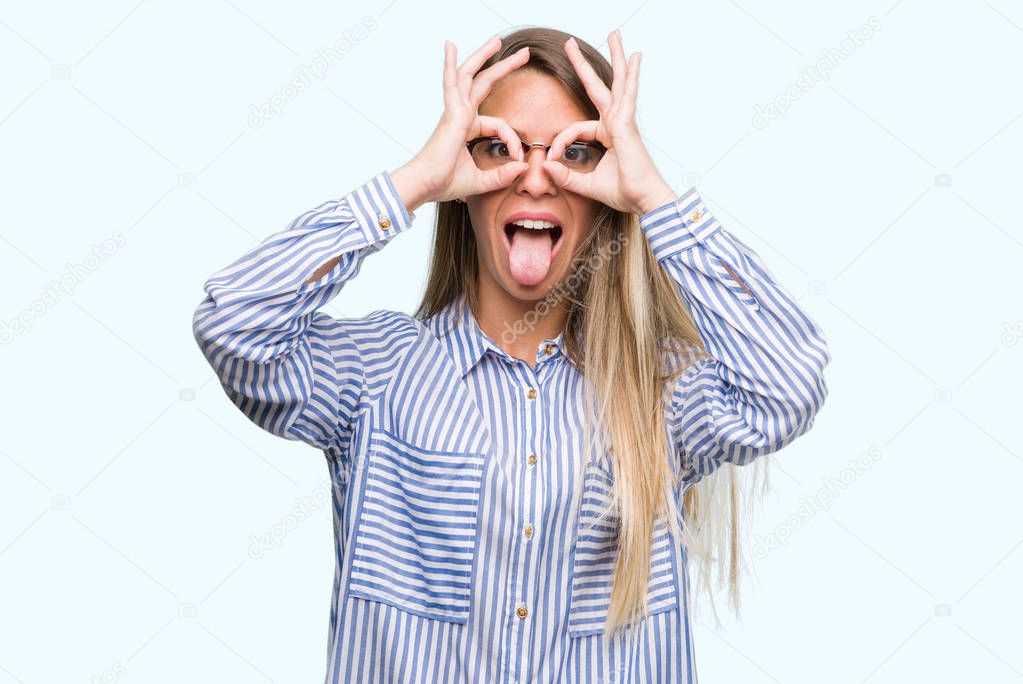 Beautiful young woman wearing elegant shirt and glasses doing ok gesture like binoculars sticking tongue out, eyes looking through fingers. Crazy expression.