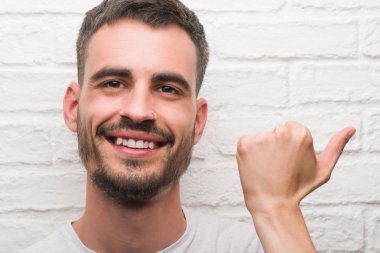 Genç yetişkin adam işaret ve başparmak kadar mutlu yüz gülümseyen tarafı ile gösterilen beyaz tuğla duvarın üzerinden ayakta