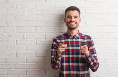 Fotoğraf makinesi ile mutlu ve komik yüz için parmakları işaret beyaz tuğla duvarın üzerinden ayakta genç yetişkin adam. İyi enerji ve vibes.