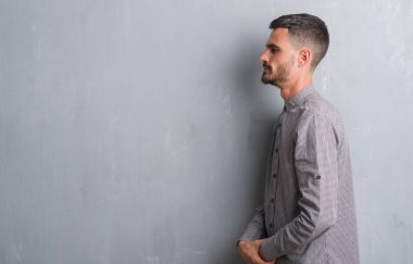 Genç yetişkin adam tarafına, seyir gri grunge duvarın üzerinden ayakta dinlenmek doğal yüz emin gülümseme ile pozuyla profili.