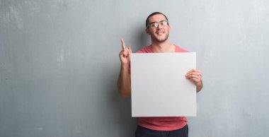 Genç beyaz adam gri grunge üzerinde boş bir afiş sürpriz bir fikir ile tutarak duvar veya gülen yüz, bir numara ile işaret parmağı soru