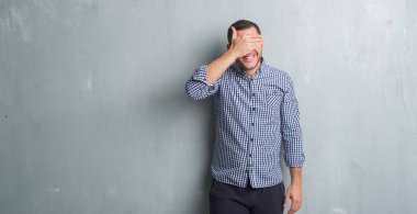 Beyaz genç gülümseyen ve gözler için sürpriz kapsayan yüz el ile gülüyor gri grunge duvarın üzerinden. Kör kavramı.