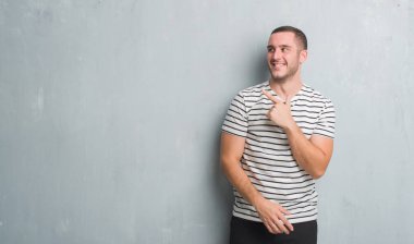 Genç beyaz adam gri grunge duvarın neşeli bir gülümseme ile el ve parmak kadar kameraya bakarak yüzündeki mutlu ve doğal ifade ile yan ile işaret yüz üzerinden.