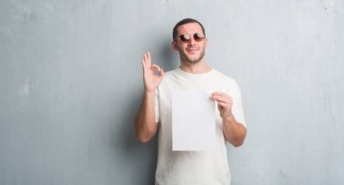 Parmak, mükemmel sembolü ile ok işareti yapıyor boş kağıt tutan gri grunge duvarın üzerinden beyaz delikanlı