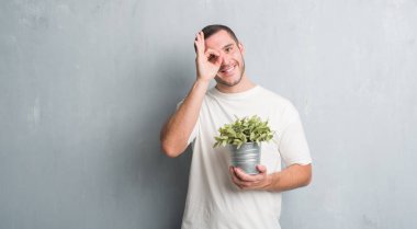 Bitki pot ile ok işareti ile el parmaklarının arasından seyir göze yapıyor gülümseyen mutlu yüz tutan gri grunge duvarın üzerinden beyaz delikanlı