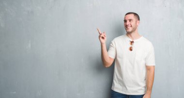 Kameraya bakarak yan el ve parmak ile işaret eden genç beyaz adam, yüzünde kocaman bir gülümseme ile güneş gözlüğü takıyor gri grunge duvarın üzerinden.