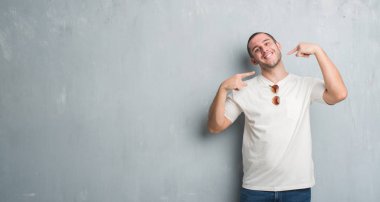 Beyaz delikanlı emin gösterilen ve işaret parmakları diş ve ağız ile gülümseyen güneş gözlüğü takıyor gri grunge duvarın üzerinden. Sağlık kavramı.