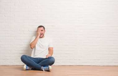 El yüz ve sürpriz duygu kendinden emin gülümseme ile tek gözüyle kapsayan beyaz tuğla duvarın üzerinden katta oturan genç beyaz adam.