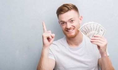 Genç Kızıl saçlı adam gri grunge holding dolar bir fikirle sürpriz duvar veya gülen yüz, bir numara ile işaret parmağı soru