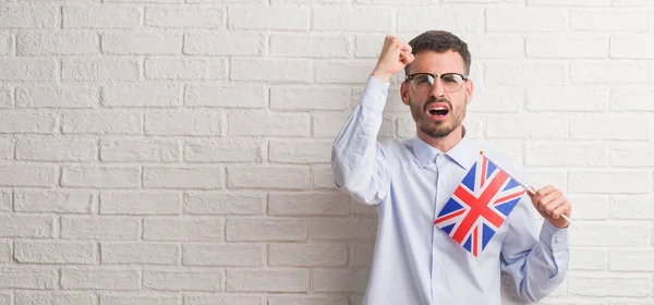 年轻的成年男子在砖墙上持有英国国旗恼怒和沮丧的呼喊与愤怒 疯狂和咆哮与举手 愤怒的概念 — 图库照片
