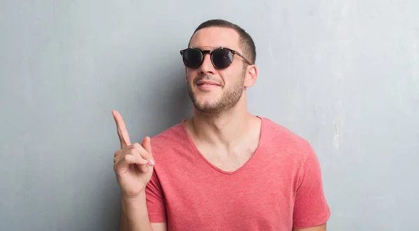 Jovem Caucasiano Sobre Parede Grunge Cinza Usando Óculos Sol Surpreso — Fotografia de Stock