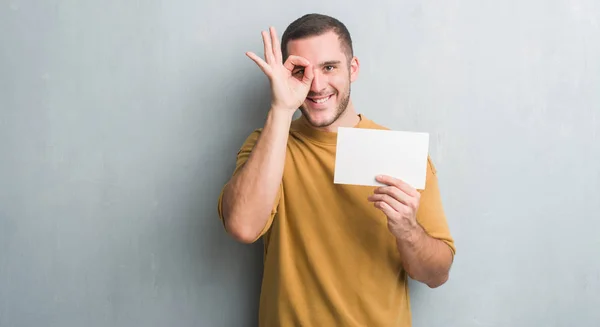 Young caucasian man over grey grunge wall holding blank card with happy face smiling doing ok sign with hand on eye looking through fingers