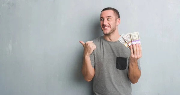 Young Caucasian Man Grey Grunge Wall Holding Bunch Money Pointing — Stock Photo, Image