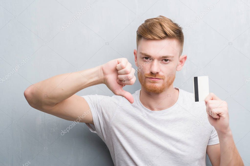 Young redhead man over grey grunge wall holding credit card with angry face, negative sign showing dislike with thumbs down, rejection concept