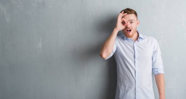 Sürpriz yüz, göz parmaklarının arasından seyir ile genç redhead iş adamı ok jest yapıyor gri grunge duvarın üzerinden şok. İmansız ifade.