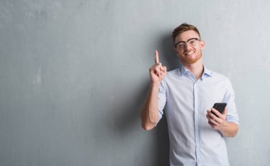Gri grunge genç Kızıl saçlı adam gönderen mesaj smartphone sürpriz bir fikir kullanarak duvar veya gülen yüz, bir numara ile işaret parmağı soru