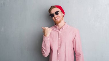 Genç Kızıl saçlı adam gri grunge duvar giyen yün kap ve işaret ve başparmak kadar mutlu yüz gülümseyen tarafı ile gösteren güneş gözlüğü