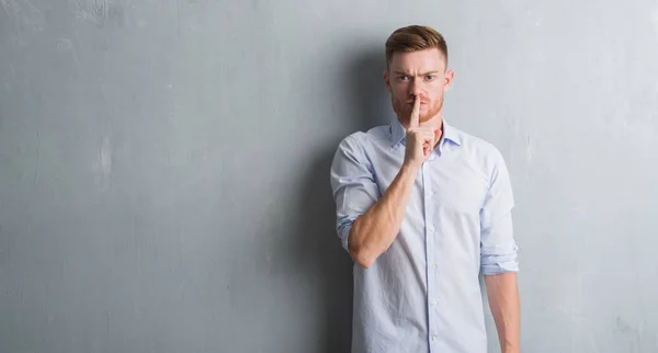 Young redhead business man over grey grunge wall asking to be quiet with finger on lips. Silence and secret concept.