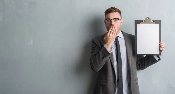 Young redhead business man over grey grunge wall holding clipboard cover mouth with hand shocked with shame for mistake, expression of fear, scared in silence, secret concept