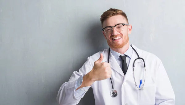 年轻的红头发的医生男人在灰色的垃圾墙快乐的大微笑做 拇指与手指 优秀的标志 — 图库照片