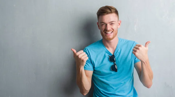 Young redhead man over grey grunge wall wearing casual outfit success sign doing positive gesture with hand, thumbs up smiling and happy. Looking at the camera with cheerful expression, winner gesture.