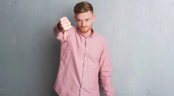 灰色垃圾墙上的年轻红衣男子穿着粉红色衬衫看起来不高兴和愤怒显示拒绝和反对的拇指向下手势 错误的表达式 — 图库照片