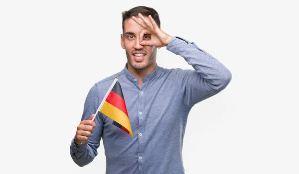 ドイツの国旗を保持して指を通して見る目に手で サインをして笑っている幸せそうな顔でハンサムな若い男 — ストック写真