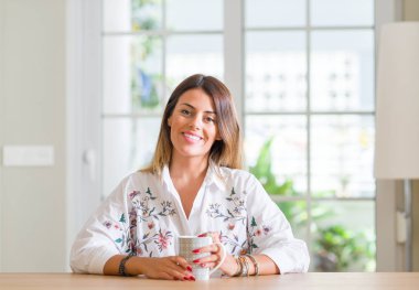 Genç kadın evde duran ve diş gösterilen kendine güvenen bir gülümsemeyle gülümseyerek mutlu bir yüz ile kahve içme