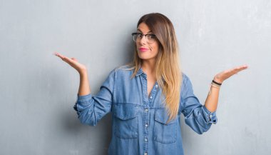 Young adult woman over grunge grey wall wearing glasses clueless and confused expression with arms and hands raised. Doubt concept. clipart