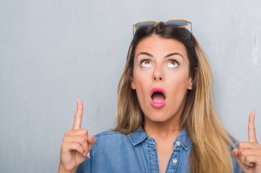 Genç Yetişkin kadın retro güneş gözlüğü takıyor grunge gri duvarın üzerinden hayran ve yukarıya bakıyor ve parmakları ile işaret ve silah kaldırdı şaşırttı.