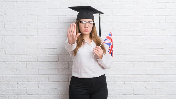 イギリスの旗を保持している大学院のキャップを身に着けている白いレンガ壁を越えて若い成人女性が防衛ジェスチャー 深刻な自信を持って式に一時停止の標識をやって手を開いてください — ストック写真