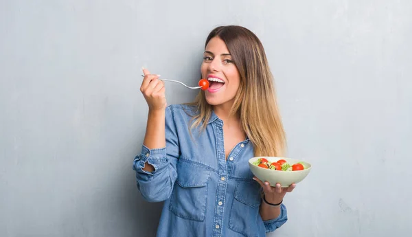 灰色垃圾墙上的年轻成年妇女吃健康的西红柿沙拉 脸上带着微笑 微笑着露出牙齿 — 图库照片