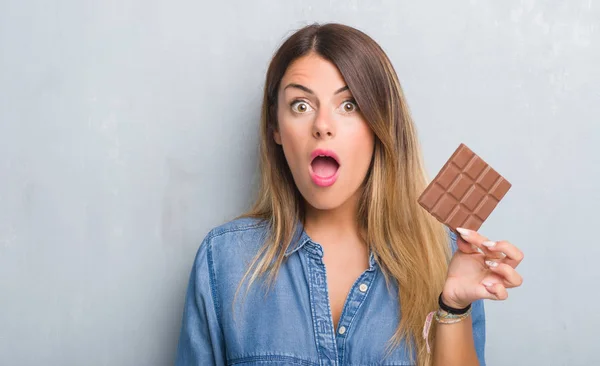 チョコレートを灰色のグランジの壁を食べる以上の若い成人女性バー驚き顔 恐怖表現と興奮とショックで怖い — ストック写真