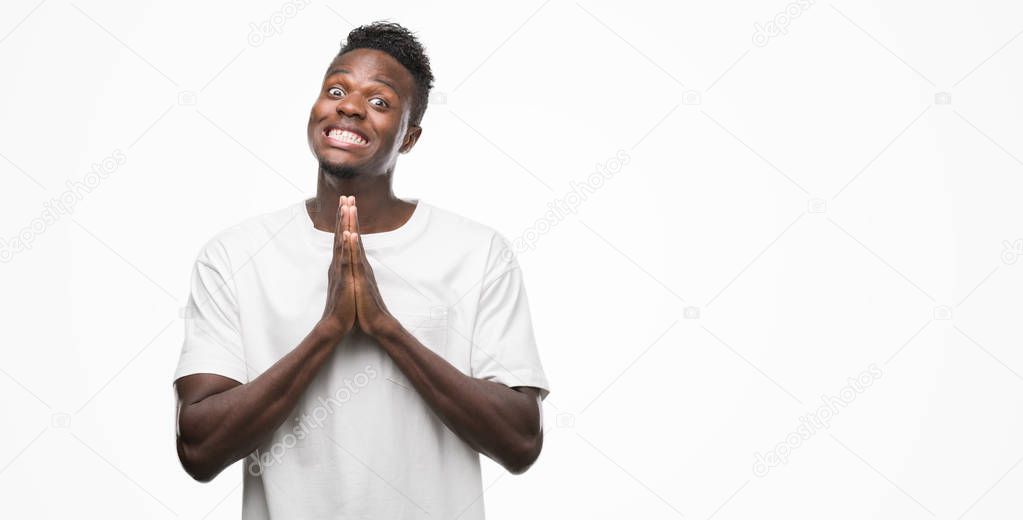 Young african american man wearing white t-shirt praying with hands together asking for forgiveness smiling confident.