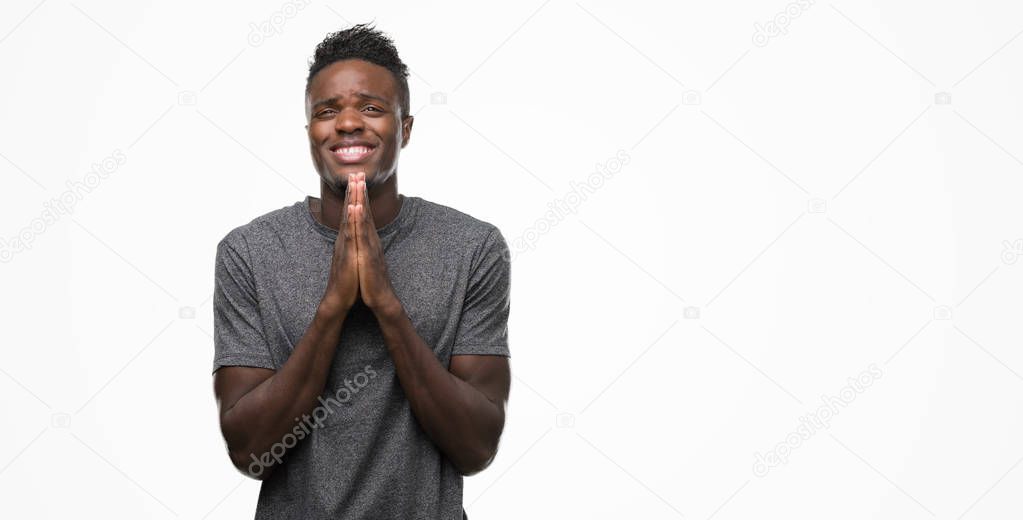 Young african american man wearing grey t-shirt praying with hands together asking for forgiveness smiling confident.