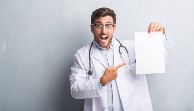 Yakışıklı genç doktor adam sayfa boş sözleşme çok mutlu el ve parmak ile işaret tutan gri grunge duvarın üzerinden