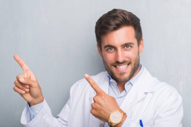 Profesyonel yakışıklı genç gülümseyen ve iki elleri ve parmakları ile tarafına işaret kamera bakıyor beyaz önlük giyen gri grunge duvarın üzerinden.