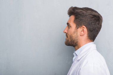 Gri grunge duvar tarafına, seyir beyaz önlük giymiş yakışıklı genç profesyonel adam rahatla profil poz ile kendine güvenen gülümseme ile doğal yüz.