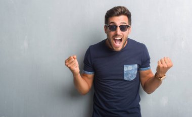 Yakışıklı delikanlı kutluyor güneş gözlüğü takıyor gri grunge duvarın üzerinden şaşırttı ve başarı için kaldırdı kollarını açarak hayret ve gözleri açık. Kazanan kavramı.