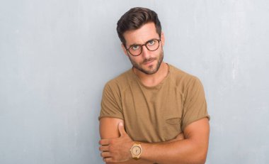 Yakışıklı delikanlı giyen gözlük şüpheci ve sinir, onaylamayan ifade yüz çapraz kolları olan gri grunge duvarın üzerinden. Negatif kişi.