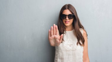 Grunge gri duvar açık eliyle dur işareti ile ciddi ve kendine güvenen ifade, savunma jest yapmak moda güneş gözlüğü takmış genç esmer kadın