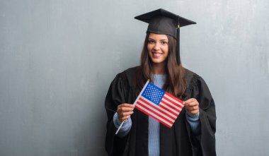 Genç esmer kadın grunge üzerinde gri bir gülen surat konumu olan Amerika'nın bayrağı tutarak ve diş gösterilen kendine güvenen bir gülümsemeyle gülümseyerek lisansüstü üniforması duvar