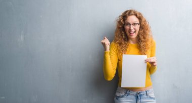 Duygu tezahürat genç Kızıl saçlı kadın gurur çığlık ve zafer ve başarı çok heyecanlı, kutluyor gri grunge duvar holding boş kağıt sayfası