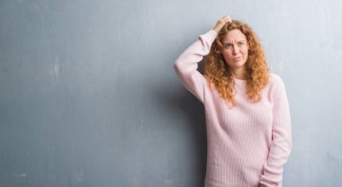 Young redhead woman over grey grunge wall wearing pink sweater confuse and wonder about question. Uncertain with doubt, thinking with hand on head. Pensive concept. clipart