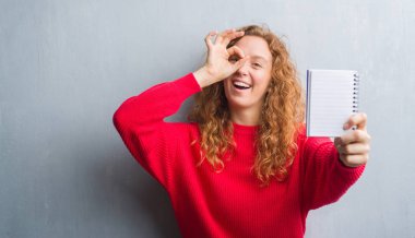 Gri grunge duvar gösteren boş defter ile mutlu yüz el parmaklarının arasından seyir göz üzerinde ok işaretiyle yapıyor gülümseyen genç Kızıl saçlı kadın