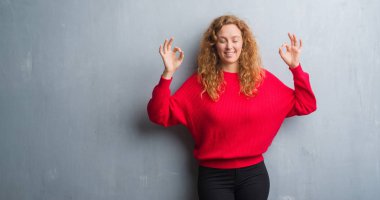 Genç Kızıl saçlı kadın duvar gri grunge giyen üzerinde kırmızı kazak rahatla ve gülümseyen gözlerle kapalı parmak jestle meditasyon yapıyor. Yoga kavramı.