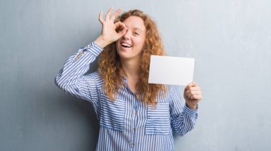 Gri grunge duvar boş kartı ile ok işareti ile el parmaklarının arasından seyir göze yapıyor gülümseyen mutlu yüz tutan genç Kızıl saçlı kadın