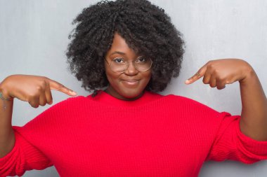 Kış kazak ve yüzünde gülümseme ile kendinden emin görünüyorsun, kendini gururlu ve mutlu parmakları ile işaret gözlük gri grunge duvarın üzerinden genç Afro-Amerikan kadın.