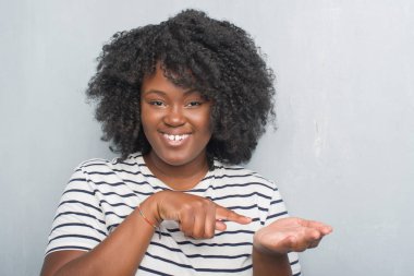 Genç Afro-Amerikan artı boyutu kadın hayrete ve el ile sunulması ve parmak ile işaret ederken kameraya gülümseyen gri grunge duvarın üzerinden.